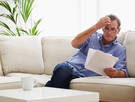 Nebenkostenabrechnung prüfen, Mann sitzt am Sofa und prüft seine Nebenkostenabrechnung, dabei fasst er sich an den Kopf, Foto: iStock/Onzeg